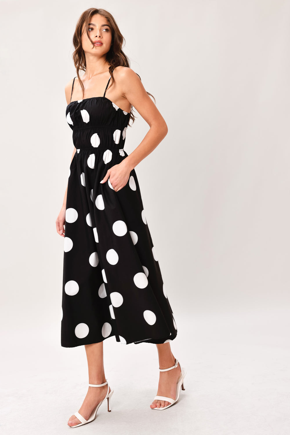 Woman wearing a black midi dress with white polka dots featuring straight neckline, straps, elastic at empire and waist, side pockets and full skirt | Flying Tomato