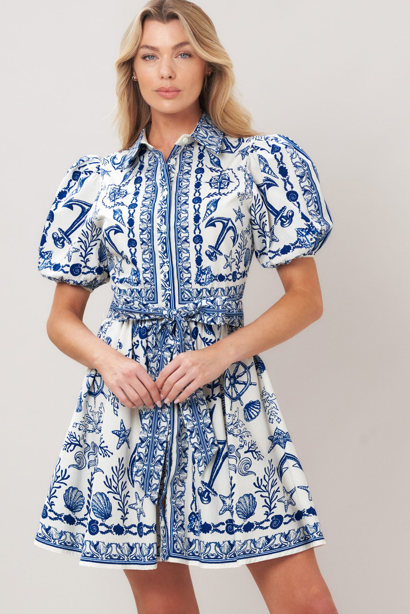 Woman wearing a blue and white patterned woven mini dress featuring collar, puff short sleeve, front button down, self sash tie and full skirt on a plain background | Flying Tomato
