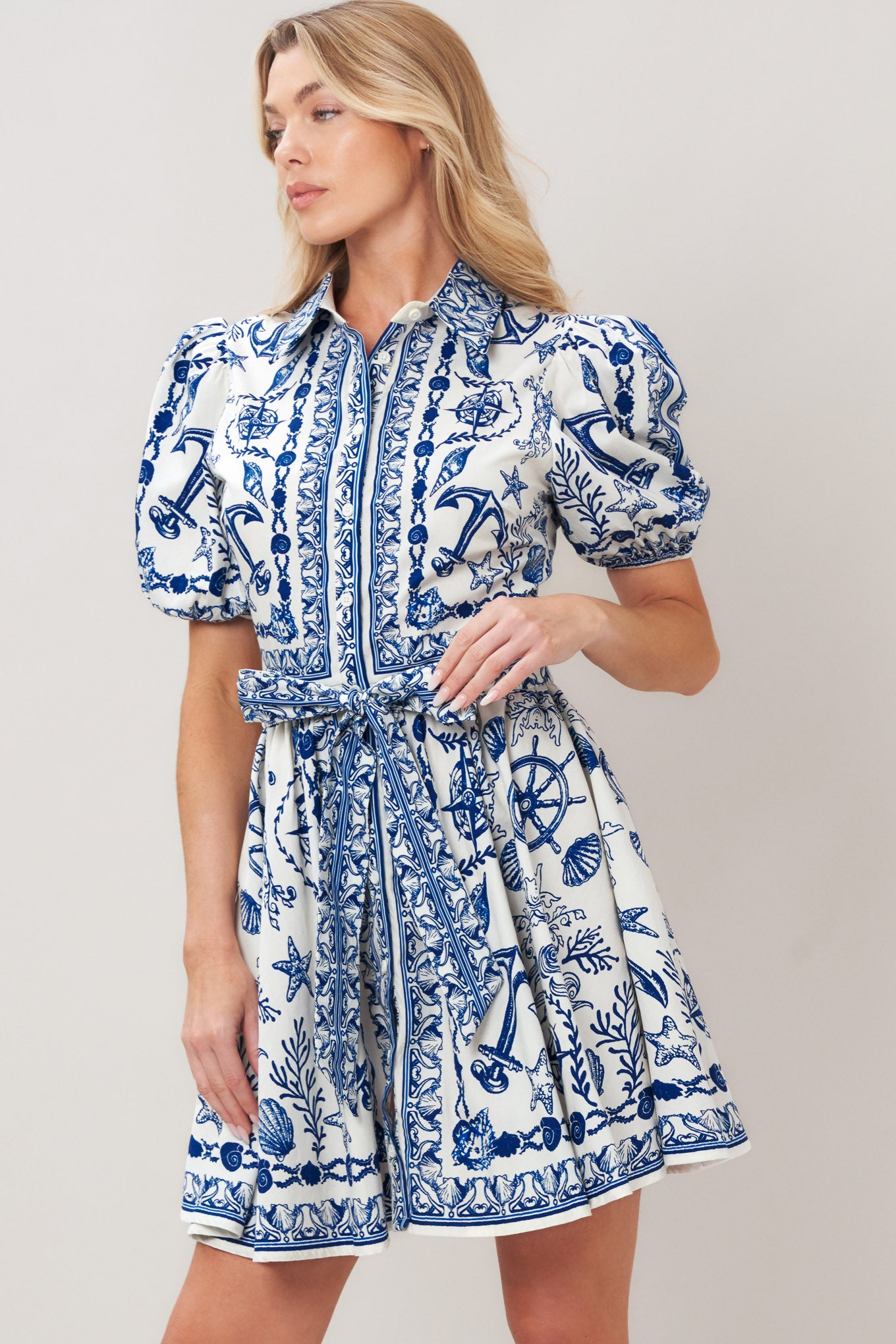 Woman wearing a blue and white patterned woven mini dress featuring collar, puff short sleeve, front button down, self sash tie and full skirt on a plain background | Flying Tomato