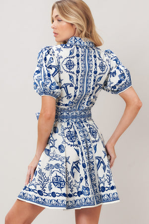 Woman wearing a blue and white patterned woven mini dress featuring collar, puff short sleeve, front button down, self sash tie and full skirt on a plain background | Flying Tomato