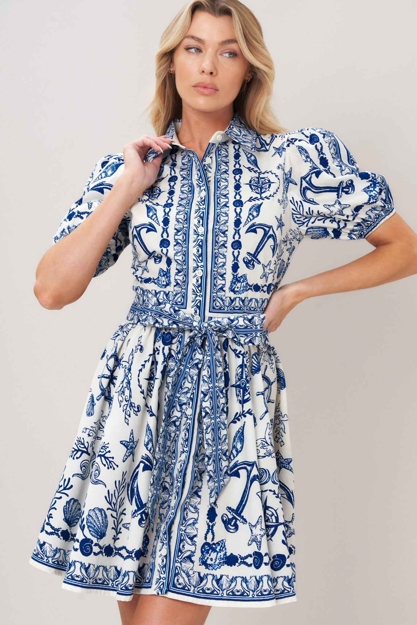 Woman wearing a blue and white patterned woven mini dress featuring collar, puff short sleeve, front button down, self sash tie and full skirt on a plain background | Flying Tomato