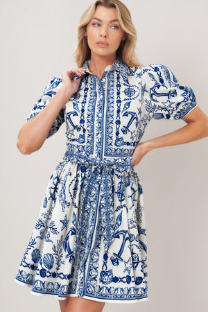 Woman wearing a blue and white patterned woven mini dress featuring collar, puff short sleeve, front button down, self sash tie and full skirt on a plain background | Flying Tomato