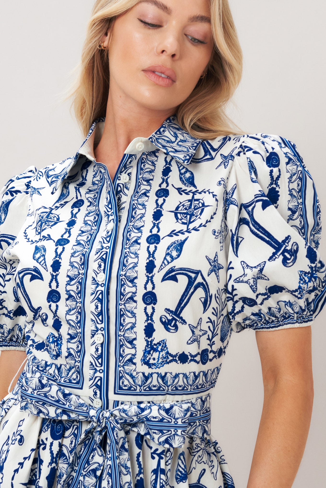 Woman wearing a blue and white patterned woven mini dress featuring collar, puff short sleeve, front button down, self sash tie and full skirt on a plain background | Flying Tomato