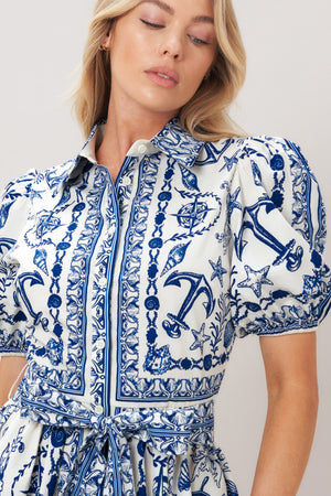 Woman wearing a blue and white patterned woven mini dress featuring collar, puff short sleeve, front button down, self sash tie and full skirt on a plain background | Flying Tomato