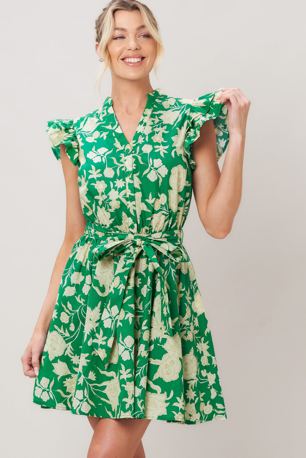 Woman wearing a green floral printed woven mini dress featuring V neckline button down, double ruffle short sleeve, elastic waist, self sash and full skirt against a plain background | Flying Tomato