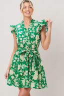 Woman wearing a green floral printed woven mini dress featuring V neckline button down, double ruffle short sleeve, elastic waist, self sash and full skirt against a plain background | Flying Tomato