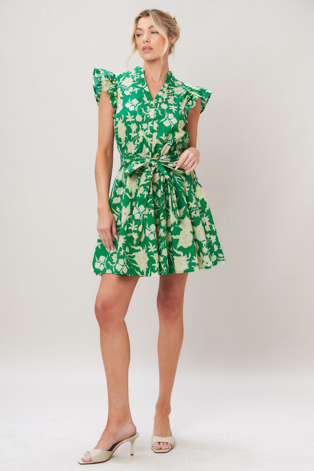 Woman wearing a green floral printed woven mini dress featuring V neckline button down, double ruffle short sleeve, elastic waist, self sash and full skirt against a plain background | Flying Tomato