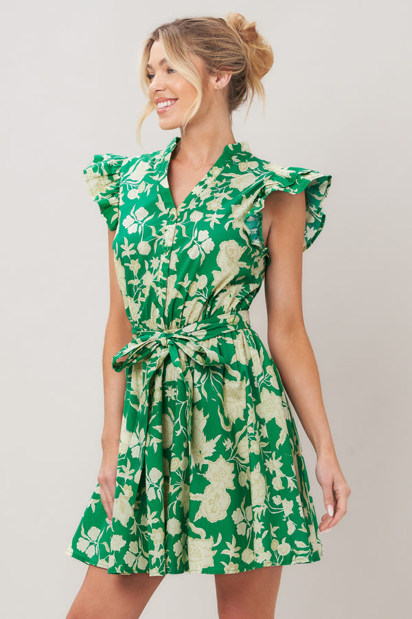 Woman wearing a green floral printed woven mini dress featuring V neckline button down, double ruffle short sleeve, elastic waist, self sash and full skirt against a plain background | Flying Tomato