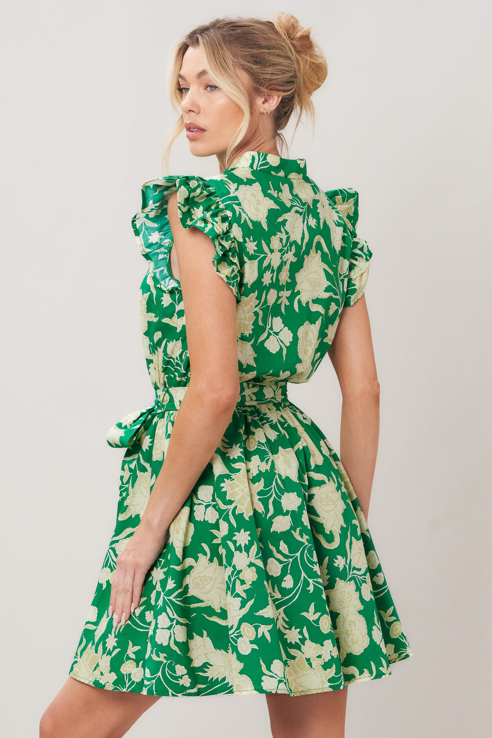 Woman wearing a green floral printed woven mini dress featuring V neckline button down, double ruffle short sleeve, elastic waist, self sash and full skirt against a plain background | Flying Tomato