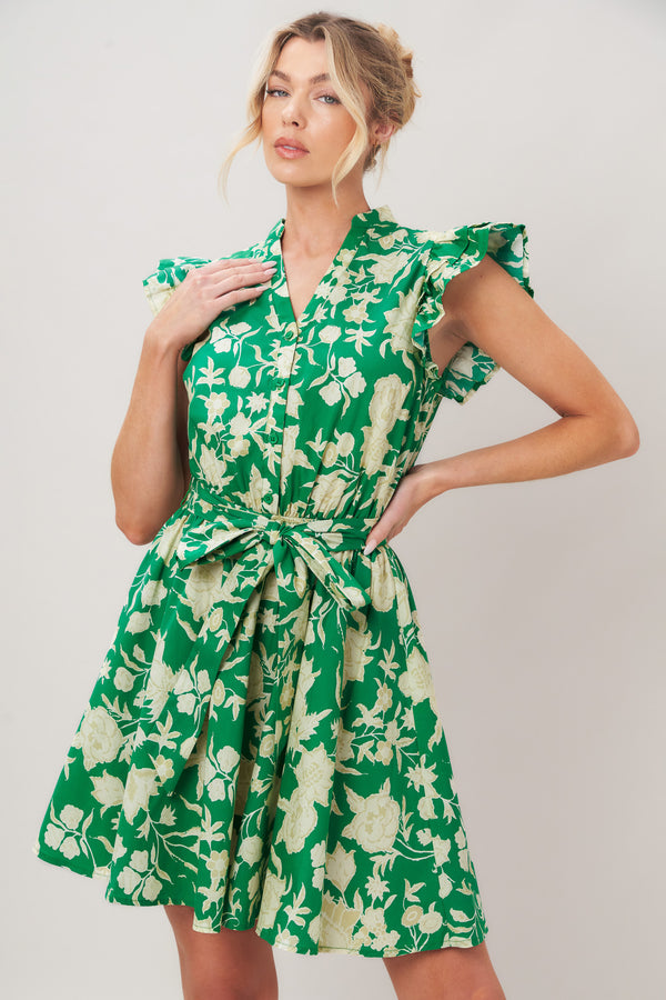 Woman wearing a green floral printed woven mini dress featuring V neckline button down, double ruffle short sleeve, elastic waist, self sash and full skirt against a plain background | Flying Tomato
