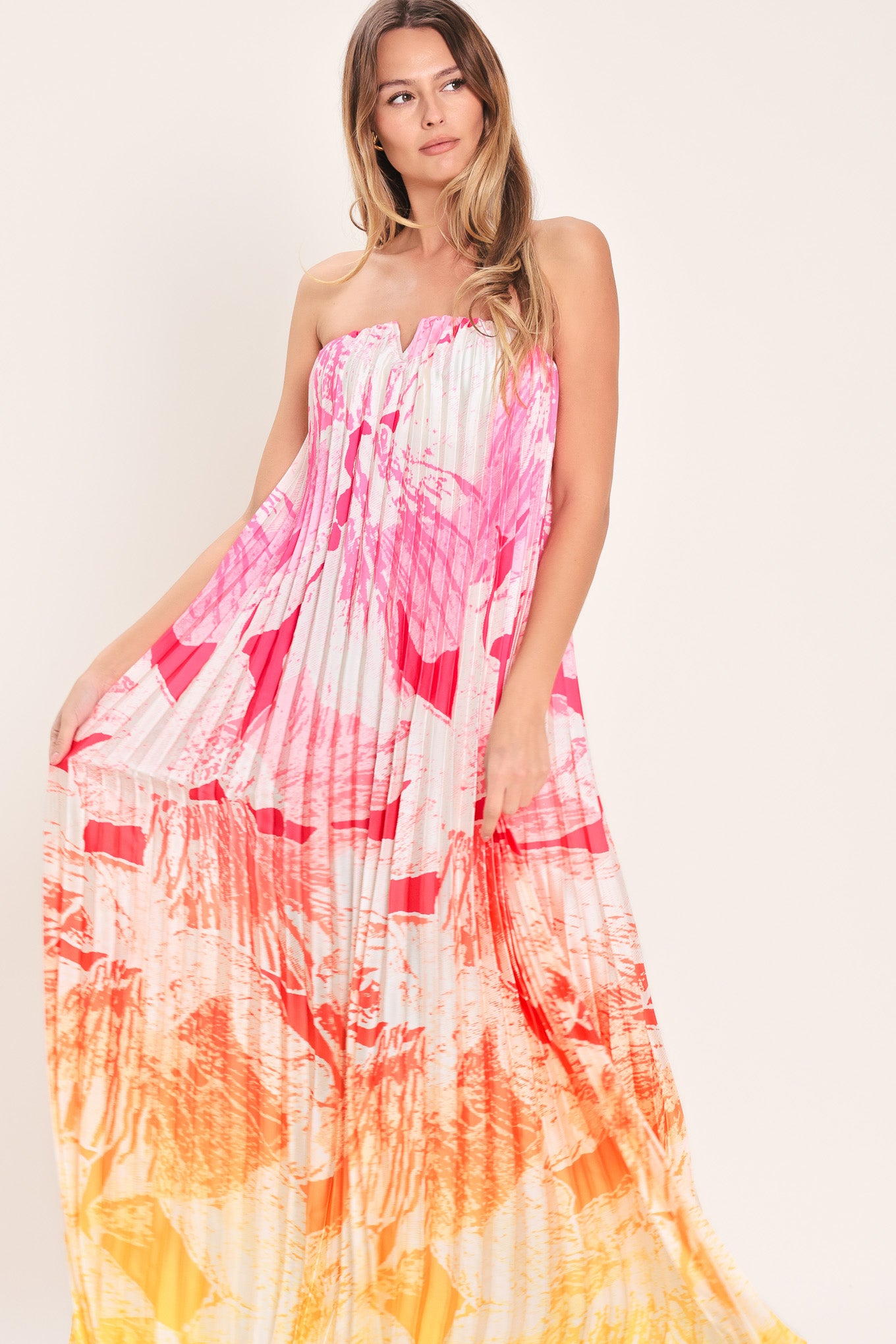 Woman wearing a pink with orange and yellow ombre pleated maxi dress with front V, strapless and back zipper closure | Flying Tomato
