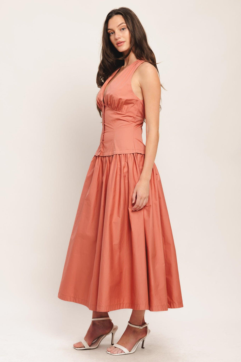 A woman models a sleeveless coral-orange midi dress with a gathered waist and pleated skirt, paired with a matching necklace and beige woven sandals, set against a warm off-white background with soft colored light leaks.