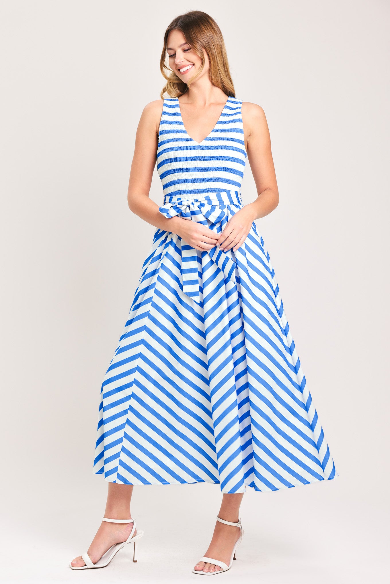Woman wearing a blue and white striped  midi dress featuring V neckline, sleeveless, smocked bodice, self sash tie and full skirt | Flying Tomato