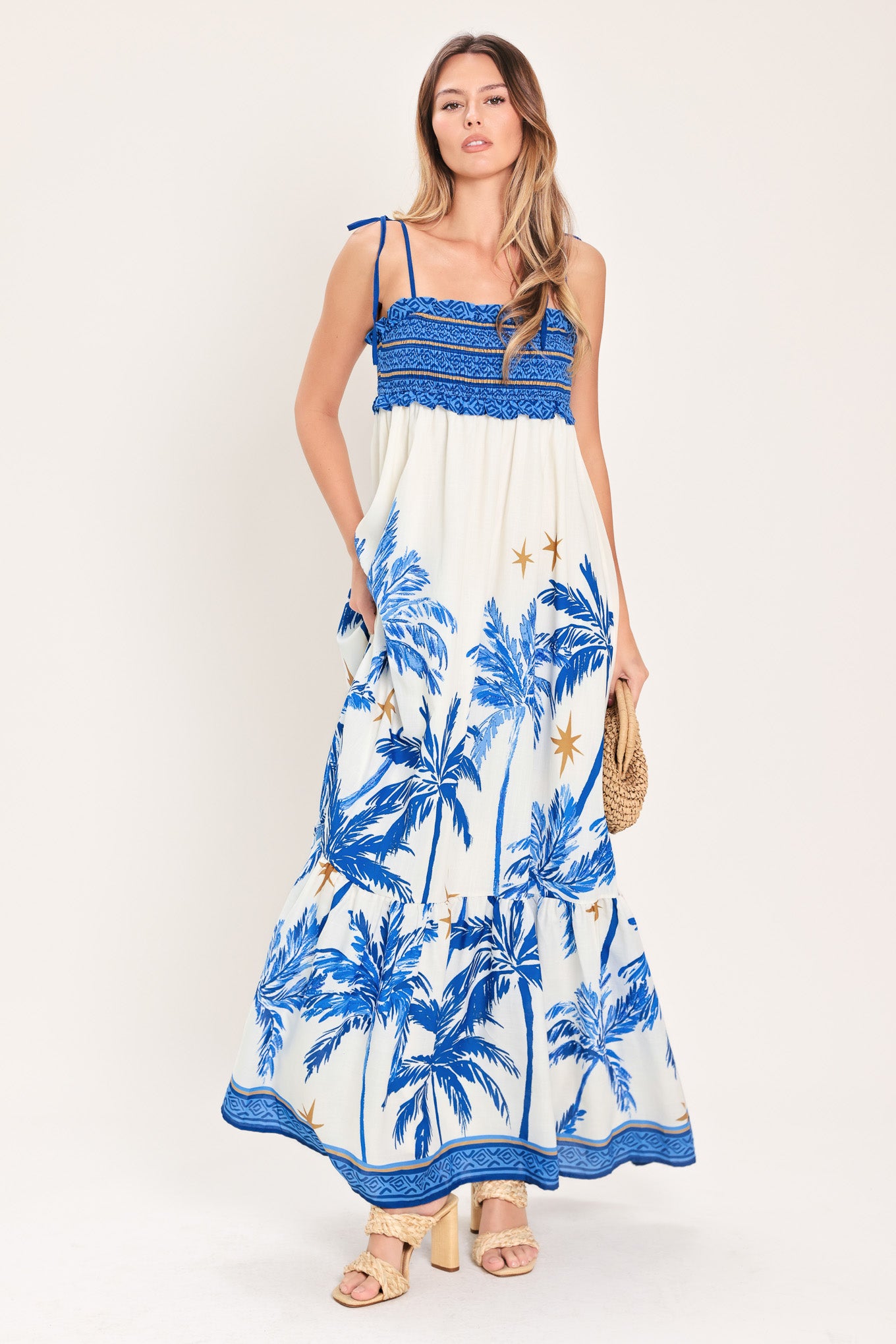 Woman wearing a blue and white palm tree patterned maxi dress featuring smocked bodice, shoulder straps, full skirt with ruffle hem | Flying Tomato