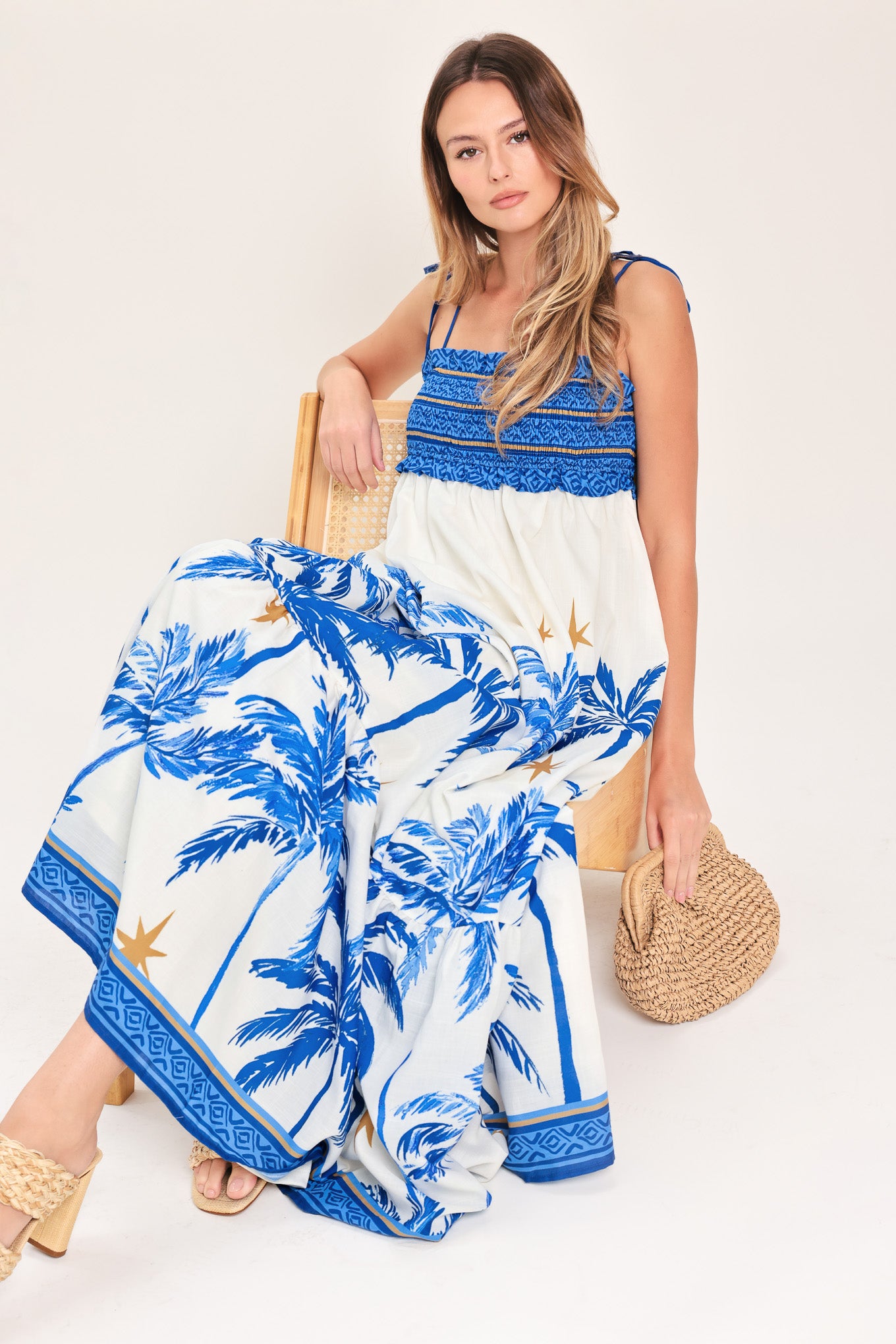 Woman wearing a blue and white palm tree patterned maxi dress featuring smocked bodice, shoulder straps, full skirt with ruffle hem | Flying Tomato