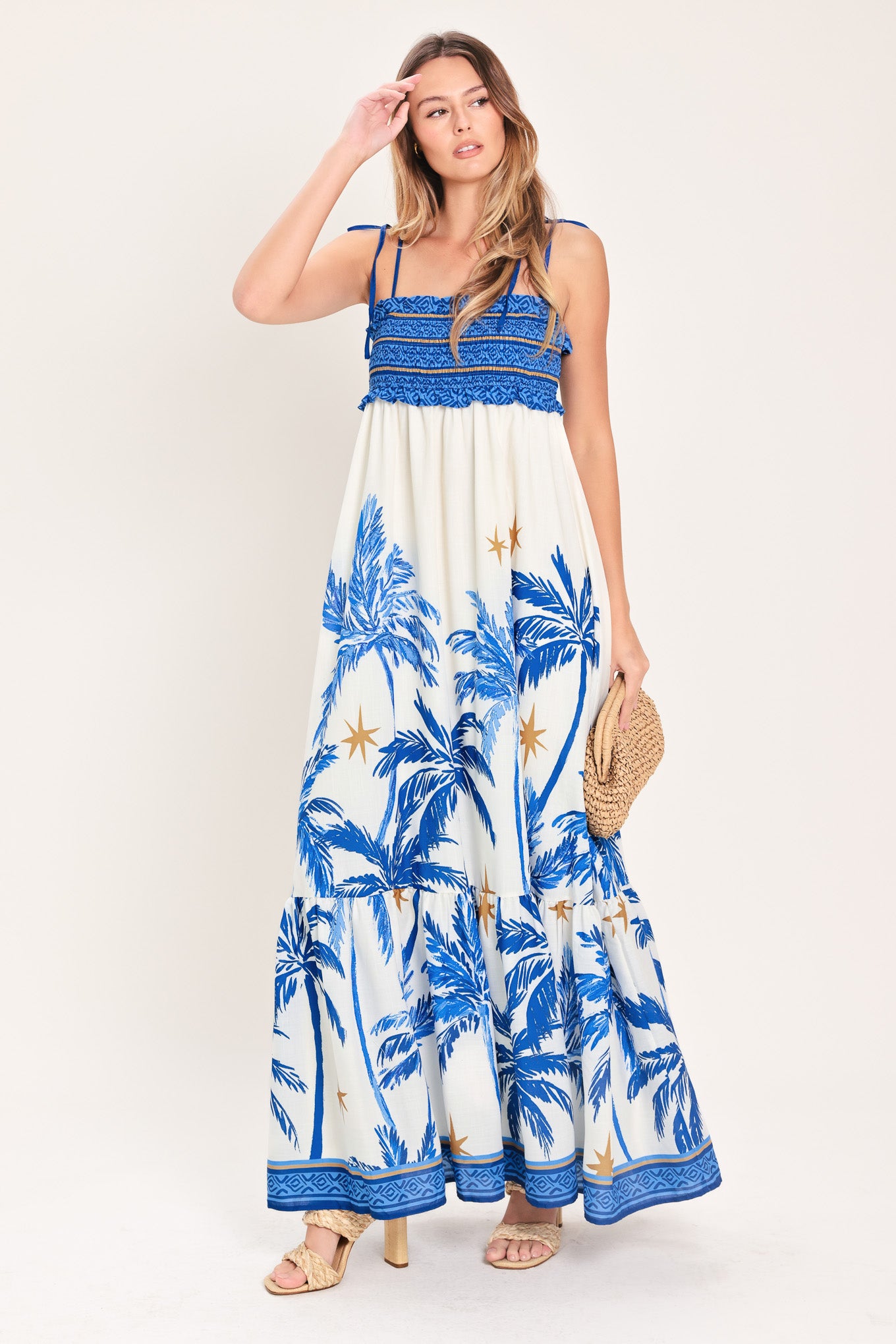 Woman wearing a blue and white palm tree patterned maxi dress featuring smocked bodice, shoulder straps, full skirt with ruffle hem | Flying Tomato