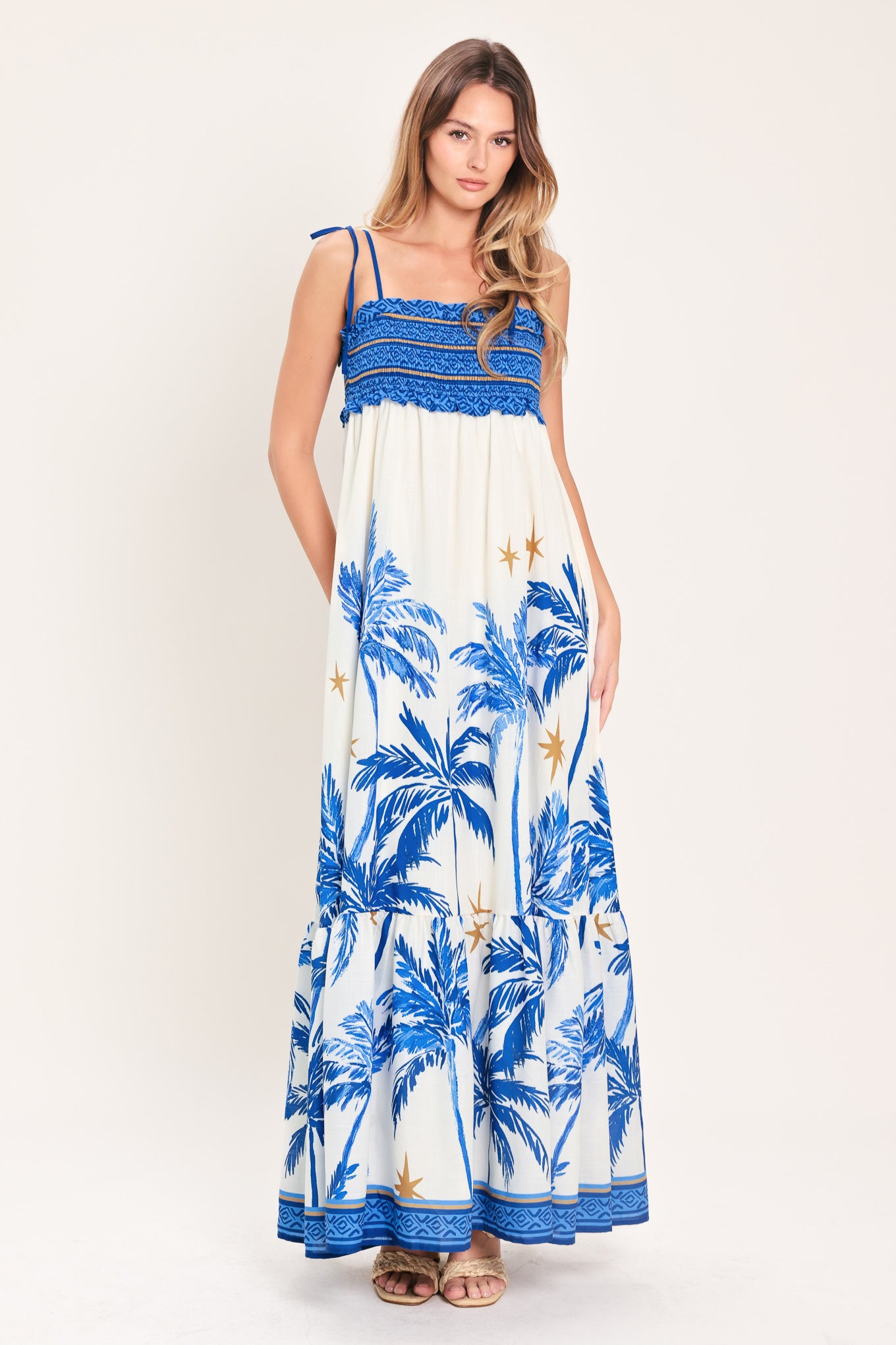 Woman wearing a blue and white palm tree patterned maxi dress featuring smocked bodice, shoulder straps, full skirt with ruffle hem | Flying Tomato