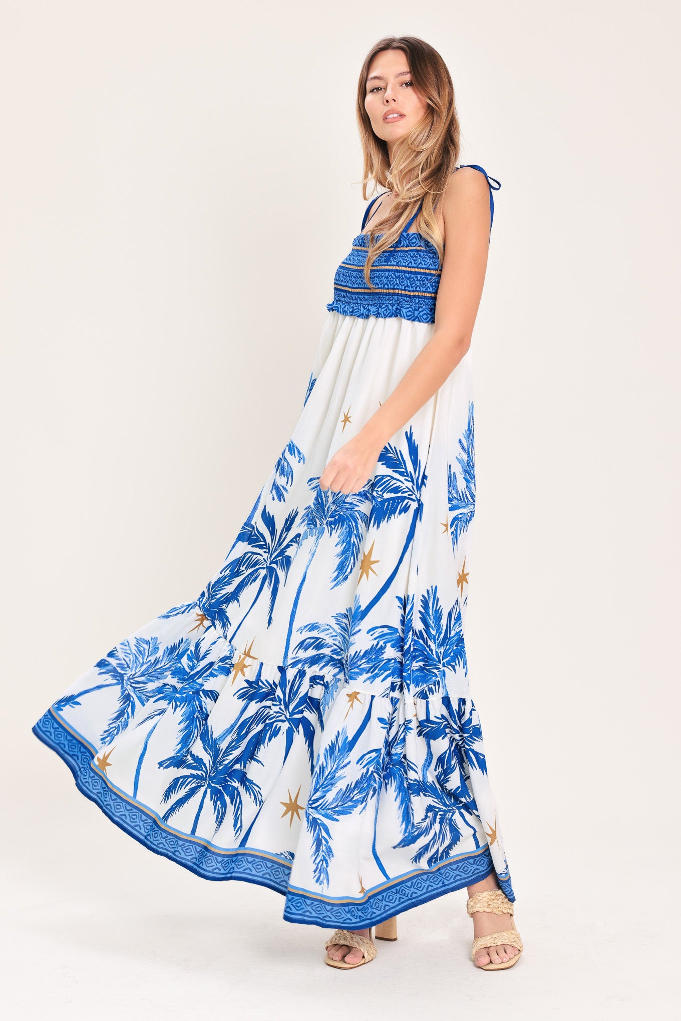 Woman wearing a blue and white palm tree patterned maxi dress featuring smocked bodice, shoulder straps, full skirt with ruffle hem | Flying Tomato