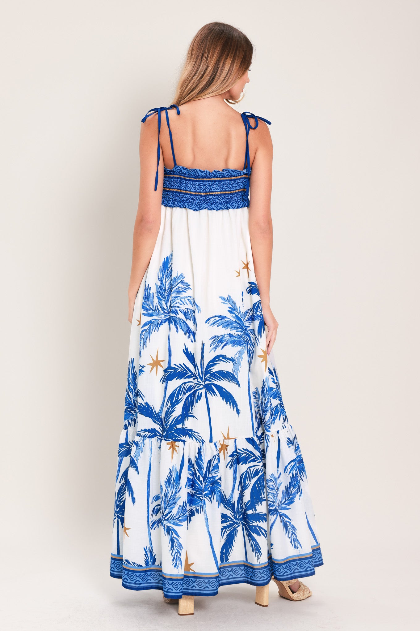 Woman wearing a blue and white palm tree patterned maxi dress featuring smocked bodice, shoulder straps, full skirt with ruffle hem | Flying Tomato