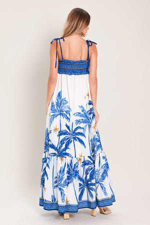 Woman wearing a blue and white palm tree patterned maxi dress featuring smocked bodice, shoulder straps, full skirt with ruffle hem | Flying Tomato