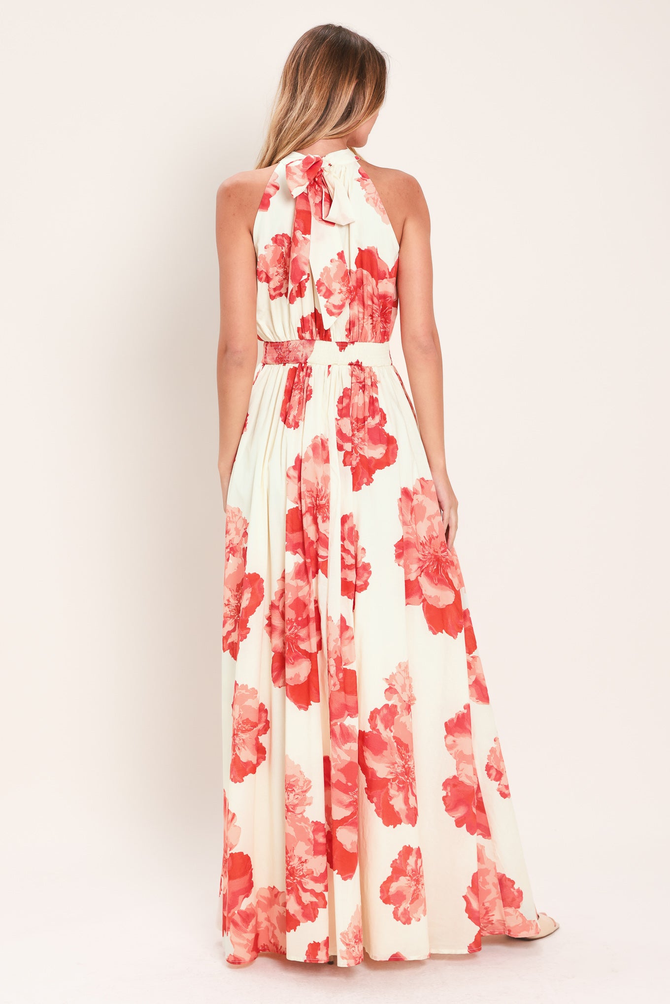 Woman wearing a cream colored with coral flowers maxi dress featuring halter neckline with back neck tie, full skirt and smocked back waistband | Flying Tomato