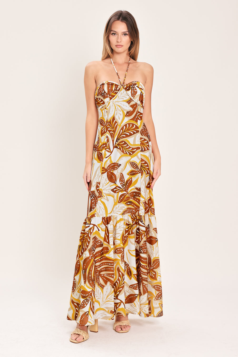 Woman wearing a mustard and brown tropical printed maxi dress featuring halter tie, ruffle hem and back zipper closure | Flying Tomato
