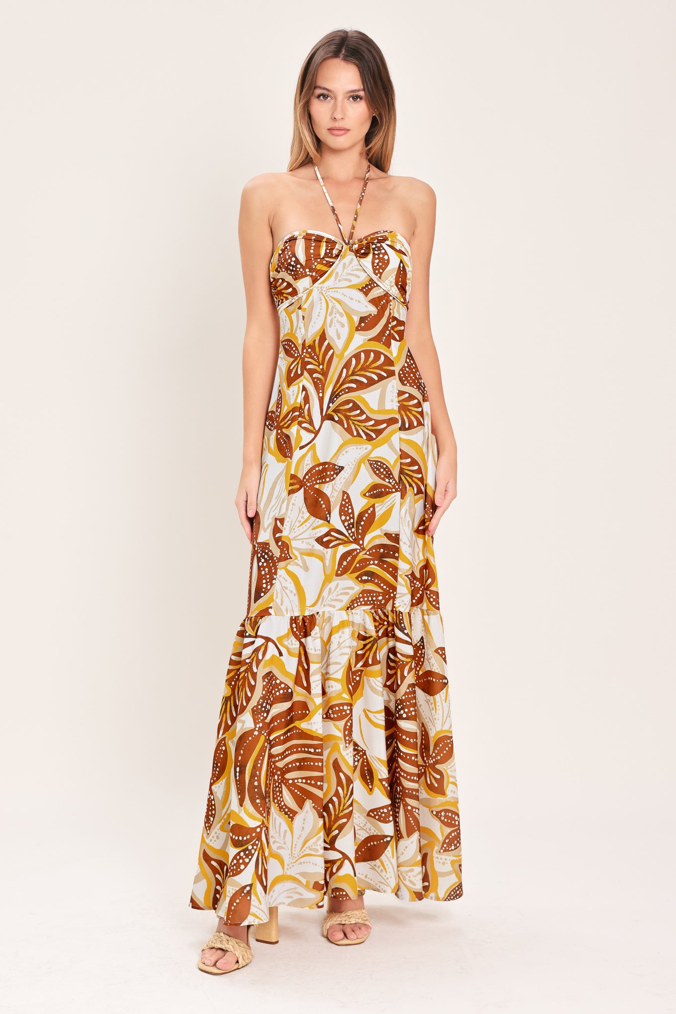 Woman wearing a mustard and brown tropical printed maxi dress featuring halter tie, ruffle hem and back zipper closure | Flying Tomato