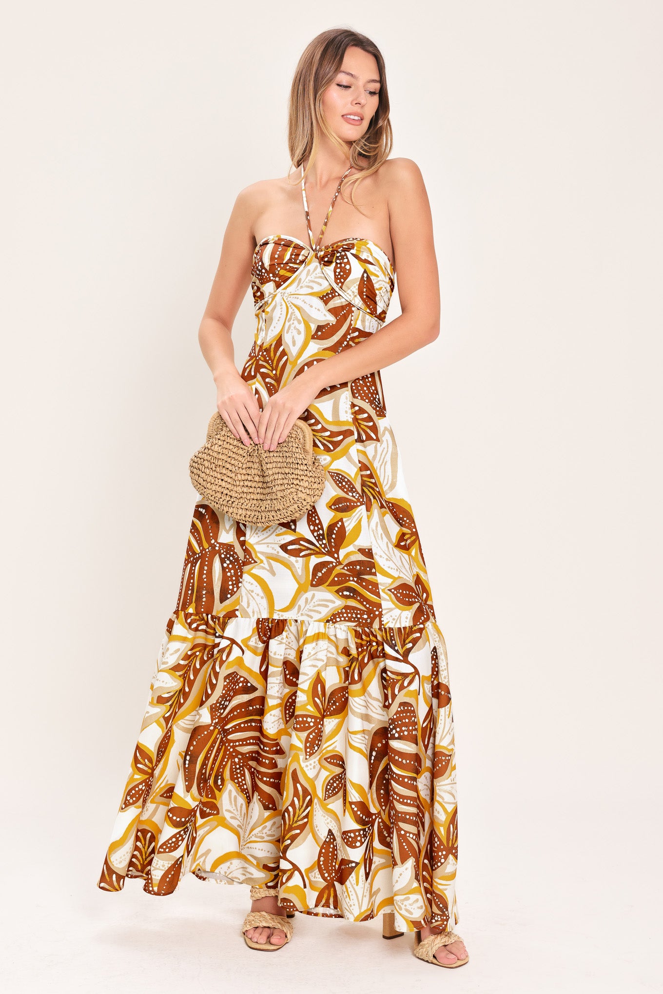 Woman wearing a mustard and brown tropical printed maxi dress featuring halter tie, ruffle hem and back zipper closure | Flying Tomato