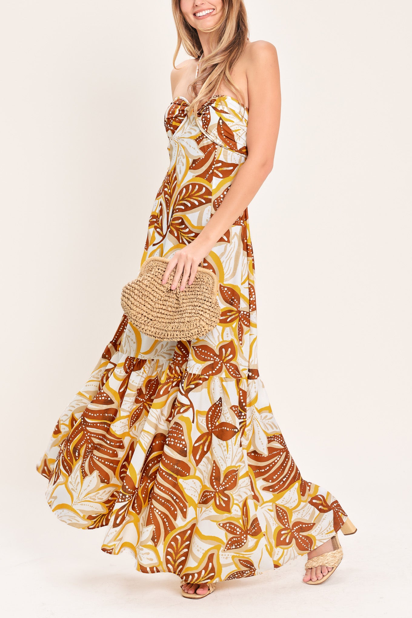 Woman wearing a mustard and brown tropical printed maxi dress featuring halter tie, ruffle hem and back zipper closure | Flying Tomato