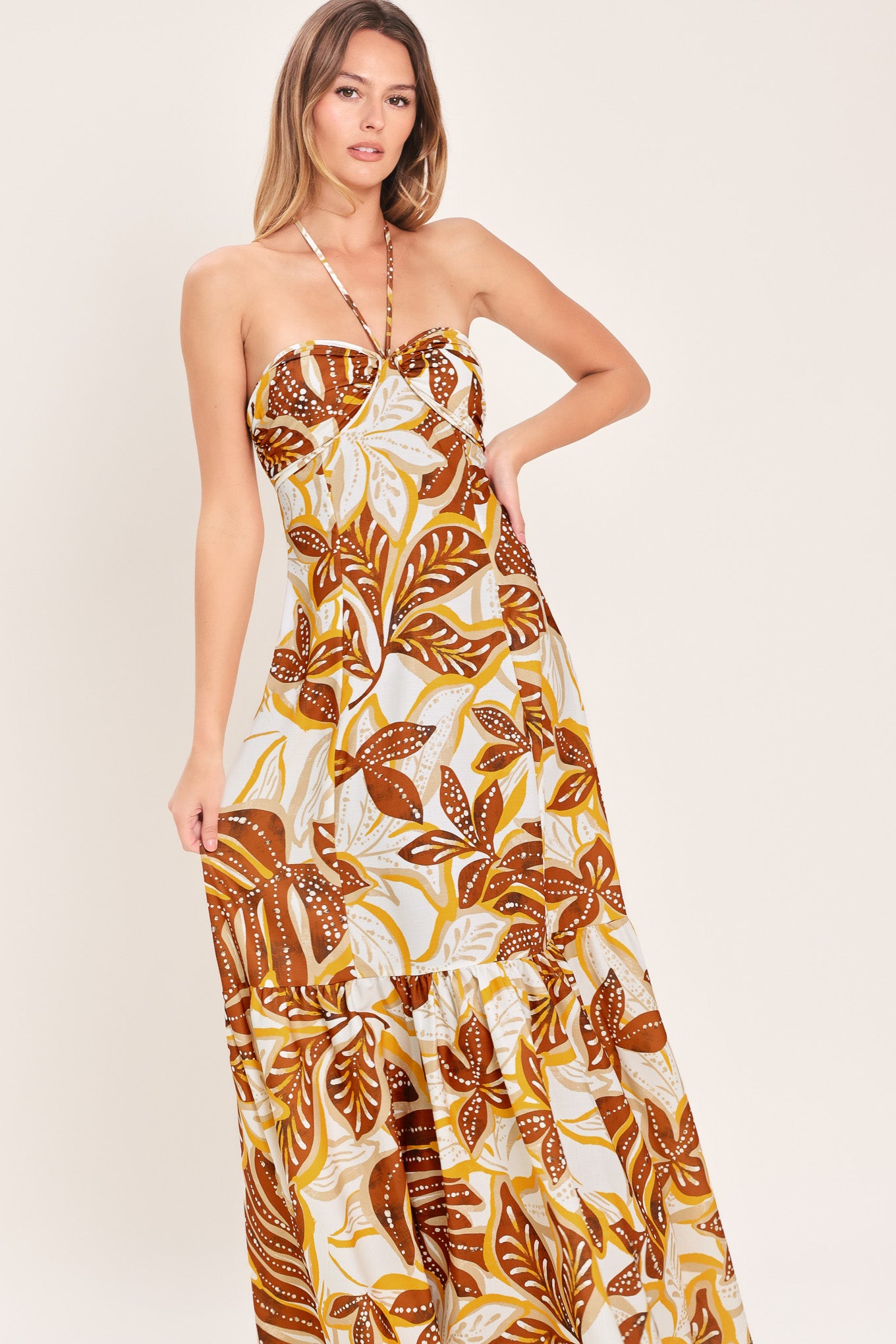 Woman wearing a mustard and brown tropical printed maxi dress featuring halter tie, ruffle hem and back zipper closure | Flying Tomato