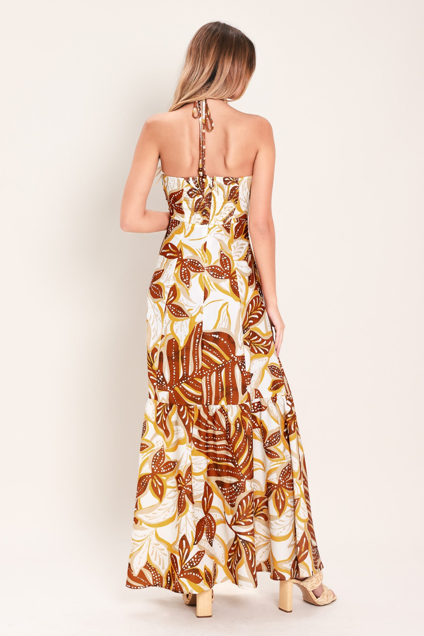 Woman wearing a mustard and brown tropical printed maxi dress featuring halter tie, ruffle hem and back zipper closure | Flying Tomato