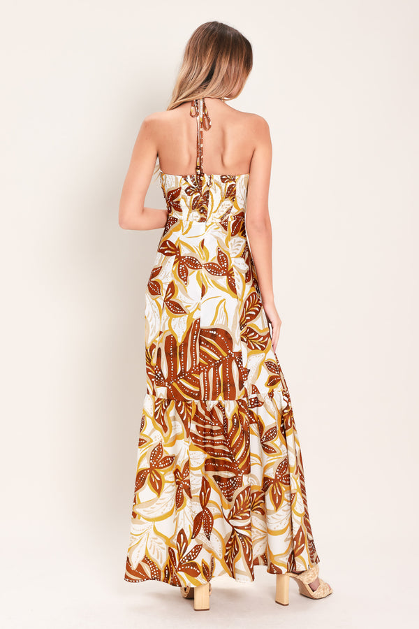 Woman wearing a mustard and brown tropical printed maxi dress featuring halter tie, ruffle hem and back zipper closure | Flying Tomato