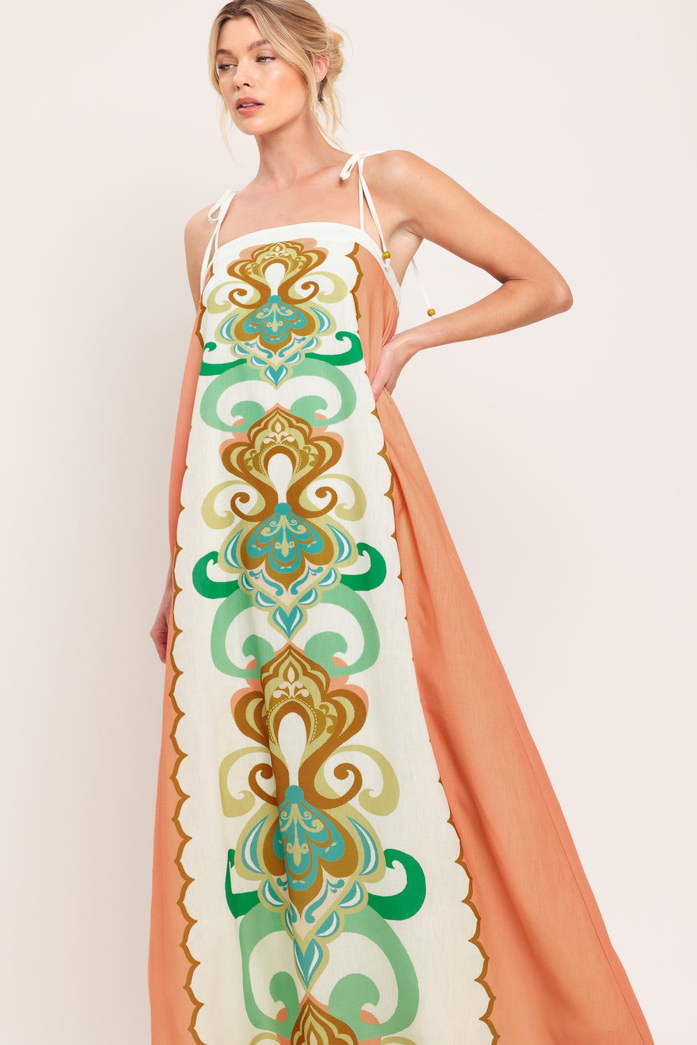 Woman wearing a long, patterned maxi dress with a peachy-brown base and green and gold design on a white background; featuring a straight neckline, shoulder ties and elastic back opening | Flying Tomato