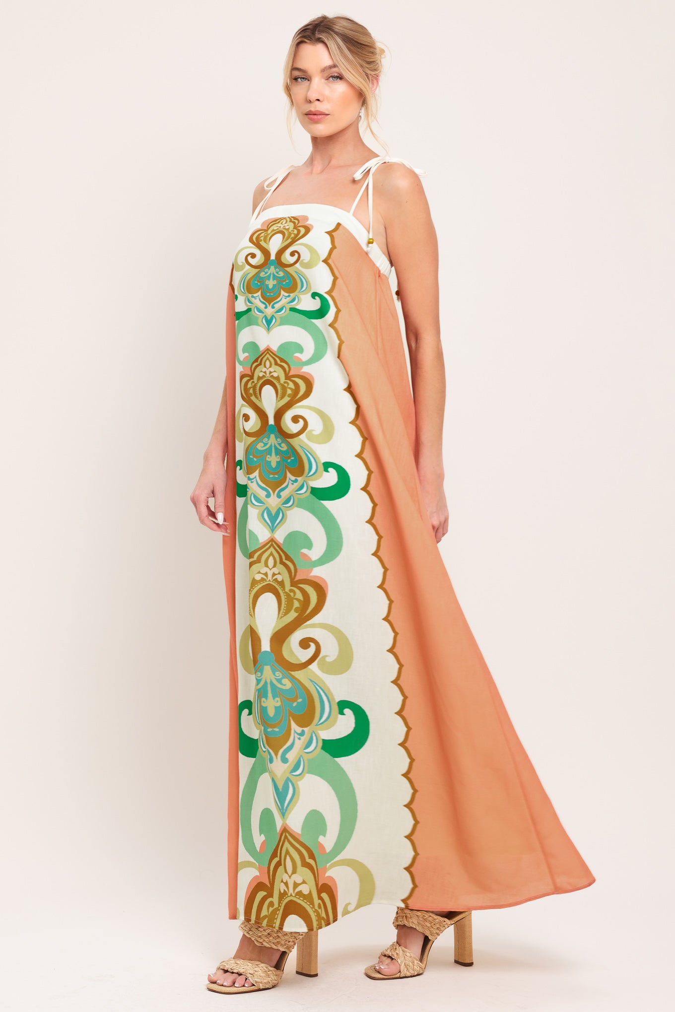 Woman wearing a long, patterned maxi dress with a peachy-brown base and green and gold design on a white background; featuring a straight neckline, shoulder ties and elastic back opening | Flying Tomato