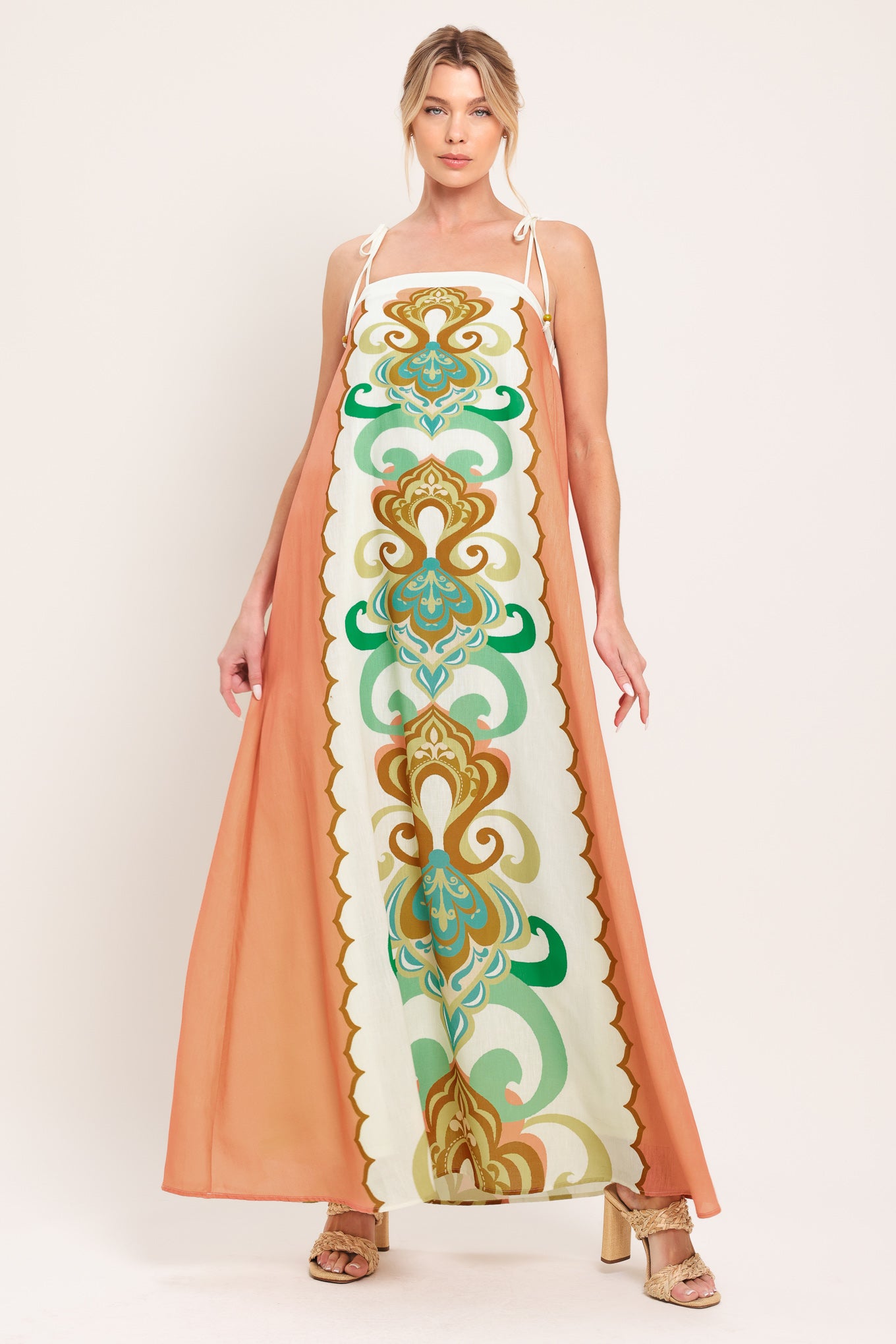Woman wearing a long, patterned maxi dress with a peachy-brown base and green and gold design on a white background; featuring a straight neckline, shoulder ties and elastic back opening | Flying Tomato