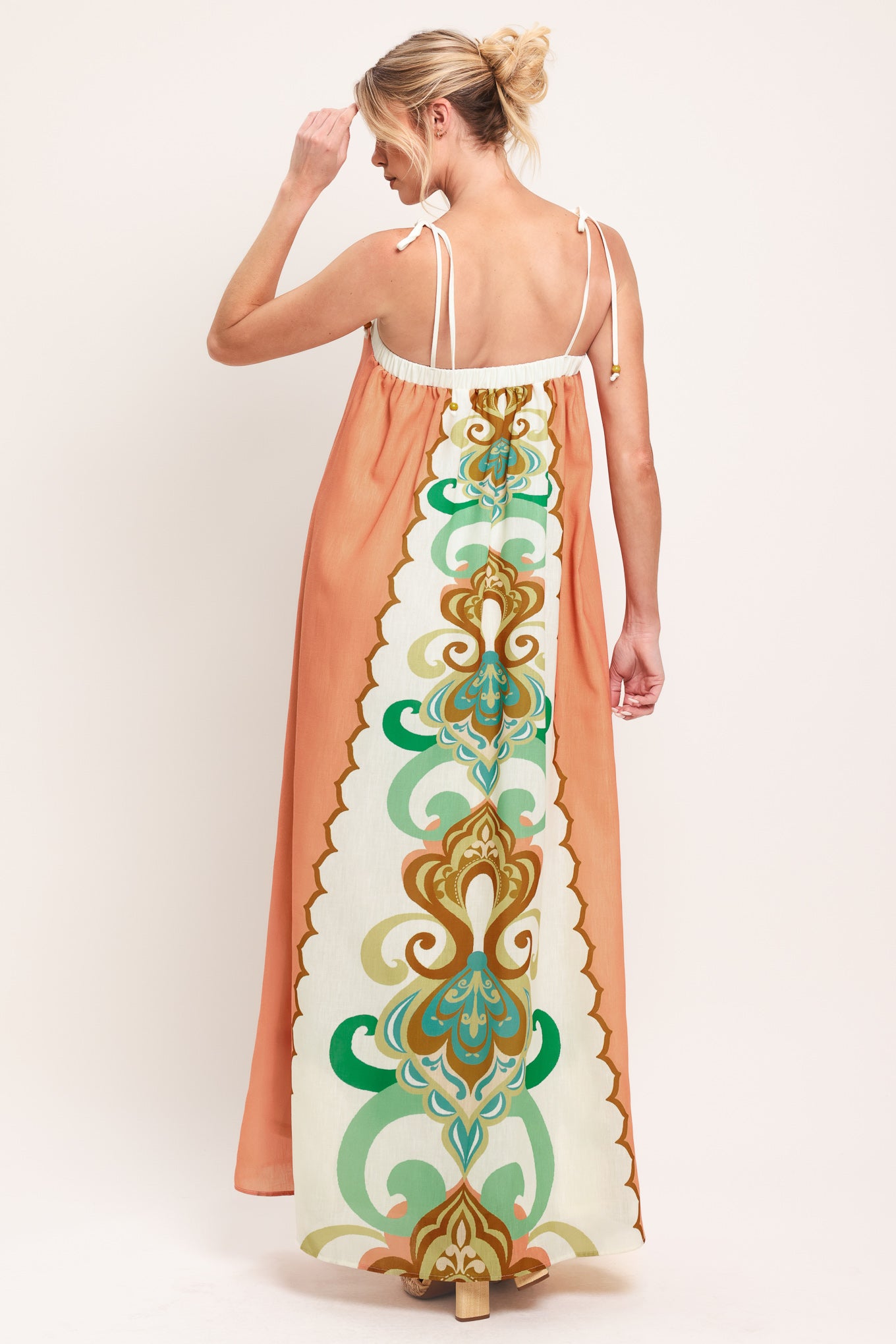 Woman wearing a long, patterned maxi dress with a peachy-brown base and green and gold design on a white background; featuring a straight neckline, shoulder ties and elastic back opening | Flying Tomato