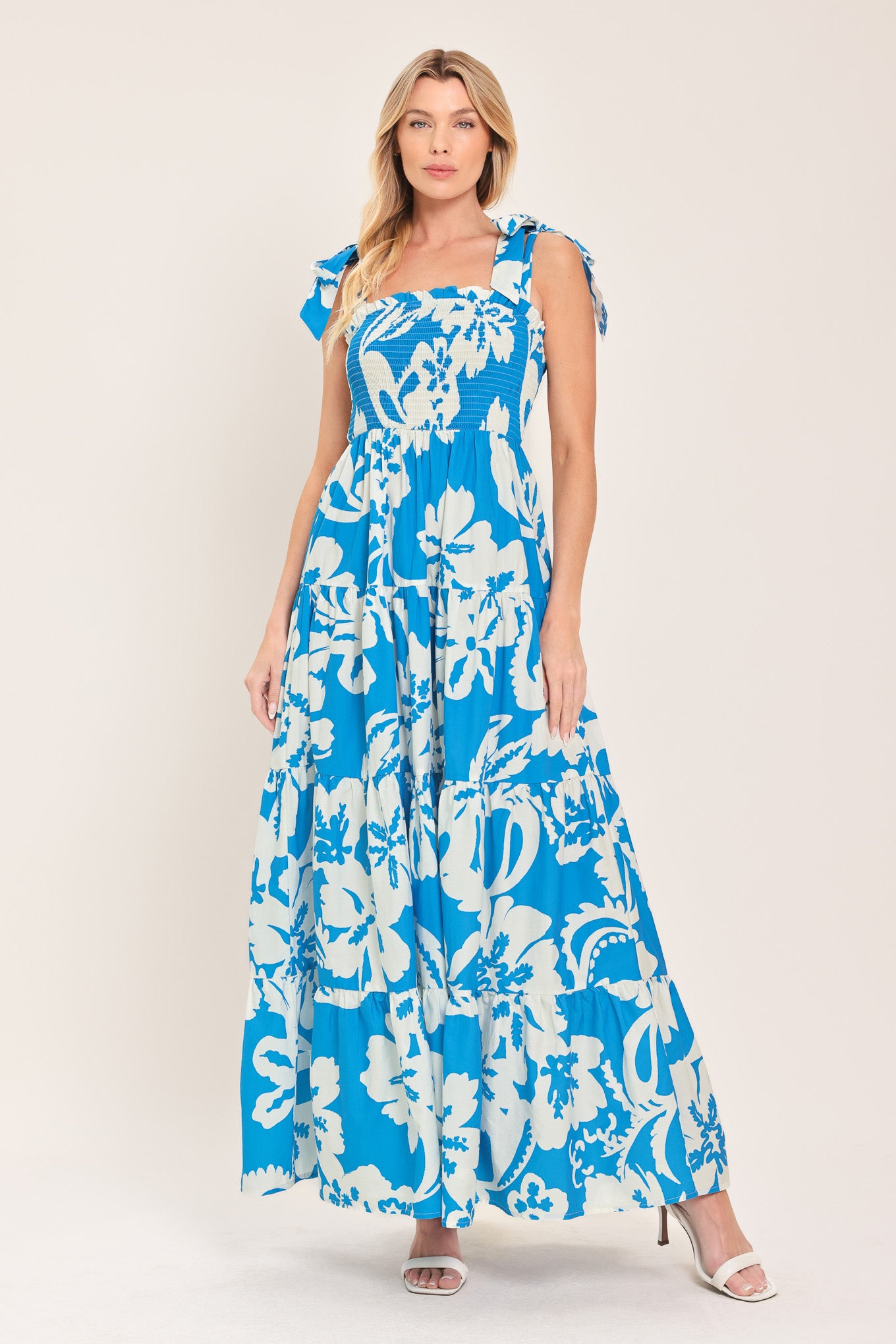 Woman wearing a blue and white tropical floral maxi dress featuring straight neckline with ruffle edge, shoulder ties, smocked bodice and tier skirt | Flying Tomato