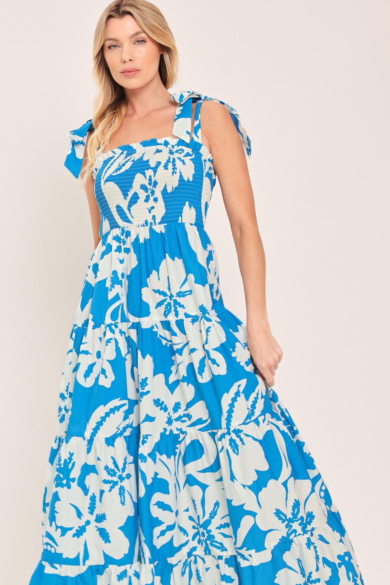 Woman wearing a blue and white tropical floral maxi dress featuring straight neckline with ruffle edge, shoulder ties, smocked bodice and tier skirt | Flying Tomato