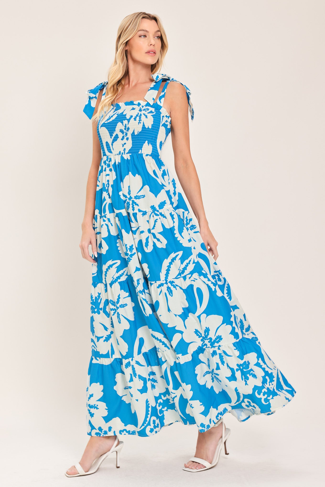 Woman wearing a blue and white tropical floral maxi dress featuring straight neckline with ruffle edge, shoulder ties, smocked bodice and tier skirt | Flying Tomato