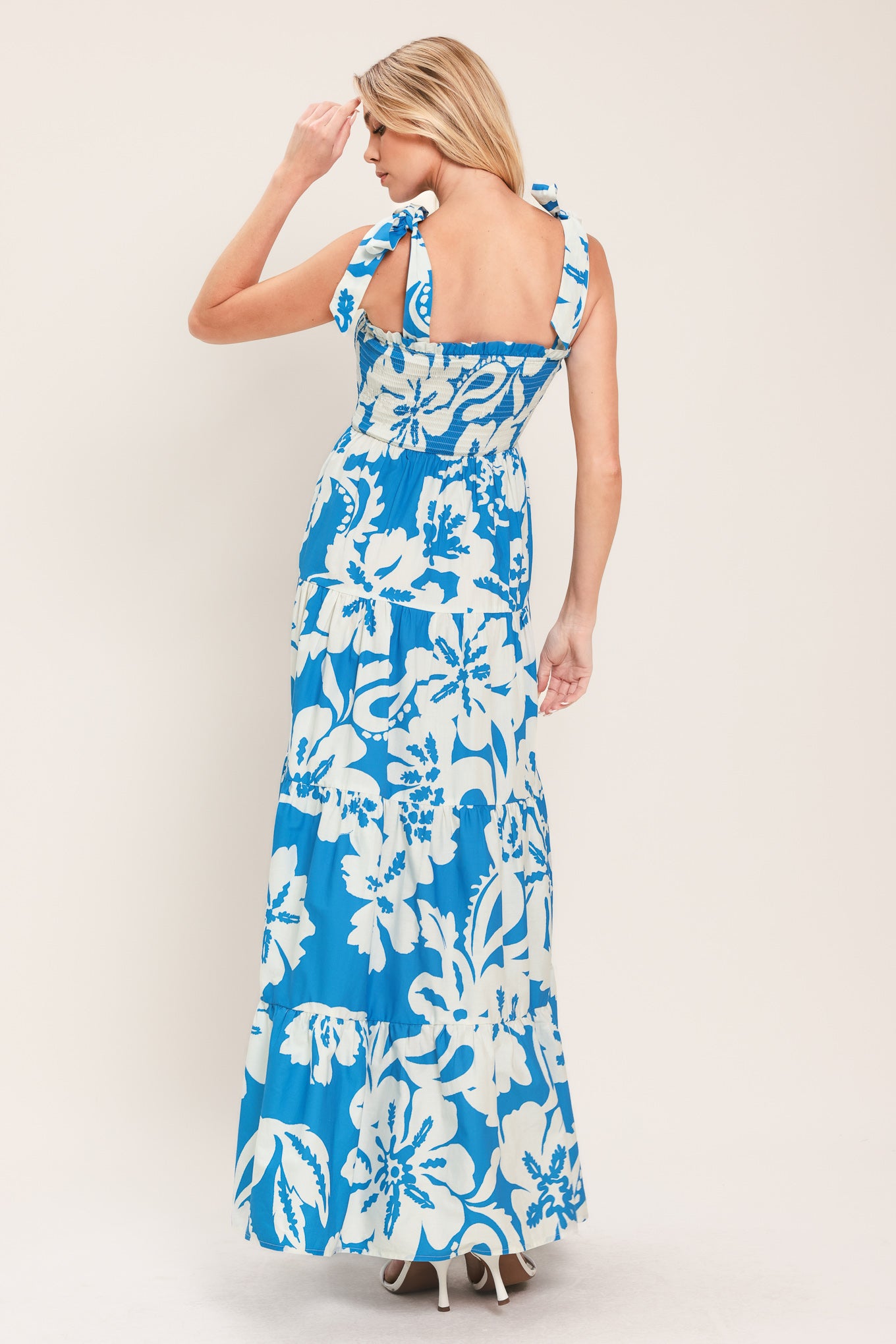 Woman wearing a blue and white tropical floral maxi dress featuring straight neckline with ruffle edge, shoulder ties, smocked bodice and tier skirt | Flying Tomato