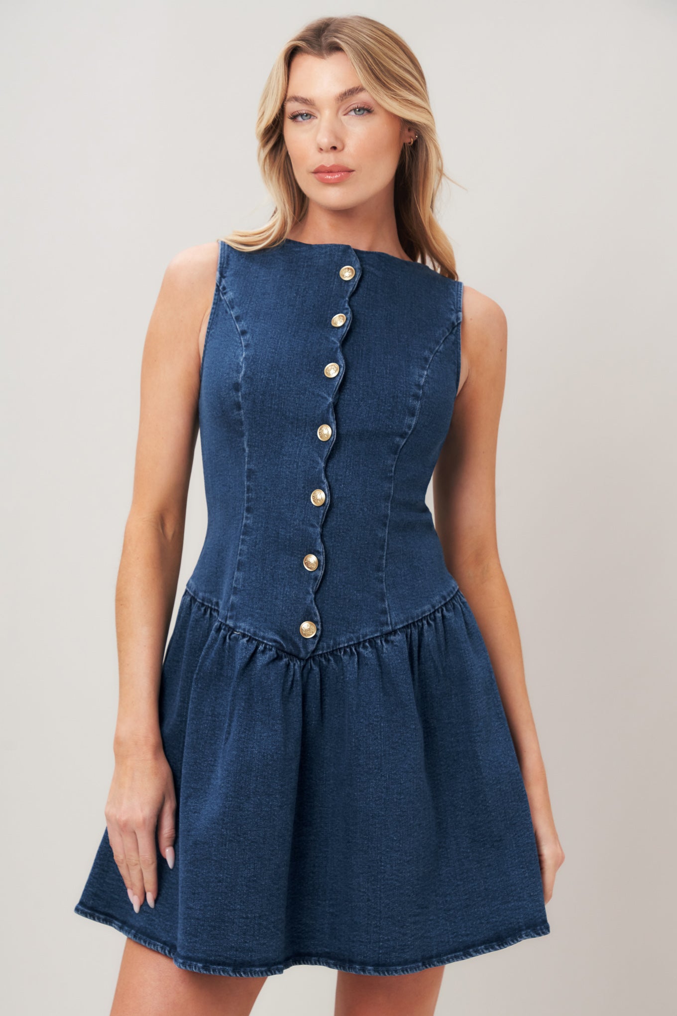 Woman wearing a blue denim mini dress featuring wide neckline, sleeveless, scalloped placket with button down detail, drop waist, full skirt and back zipper closure on a plain background | Flying Tomato
