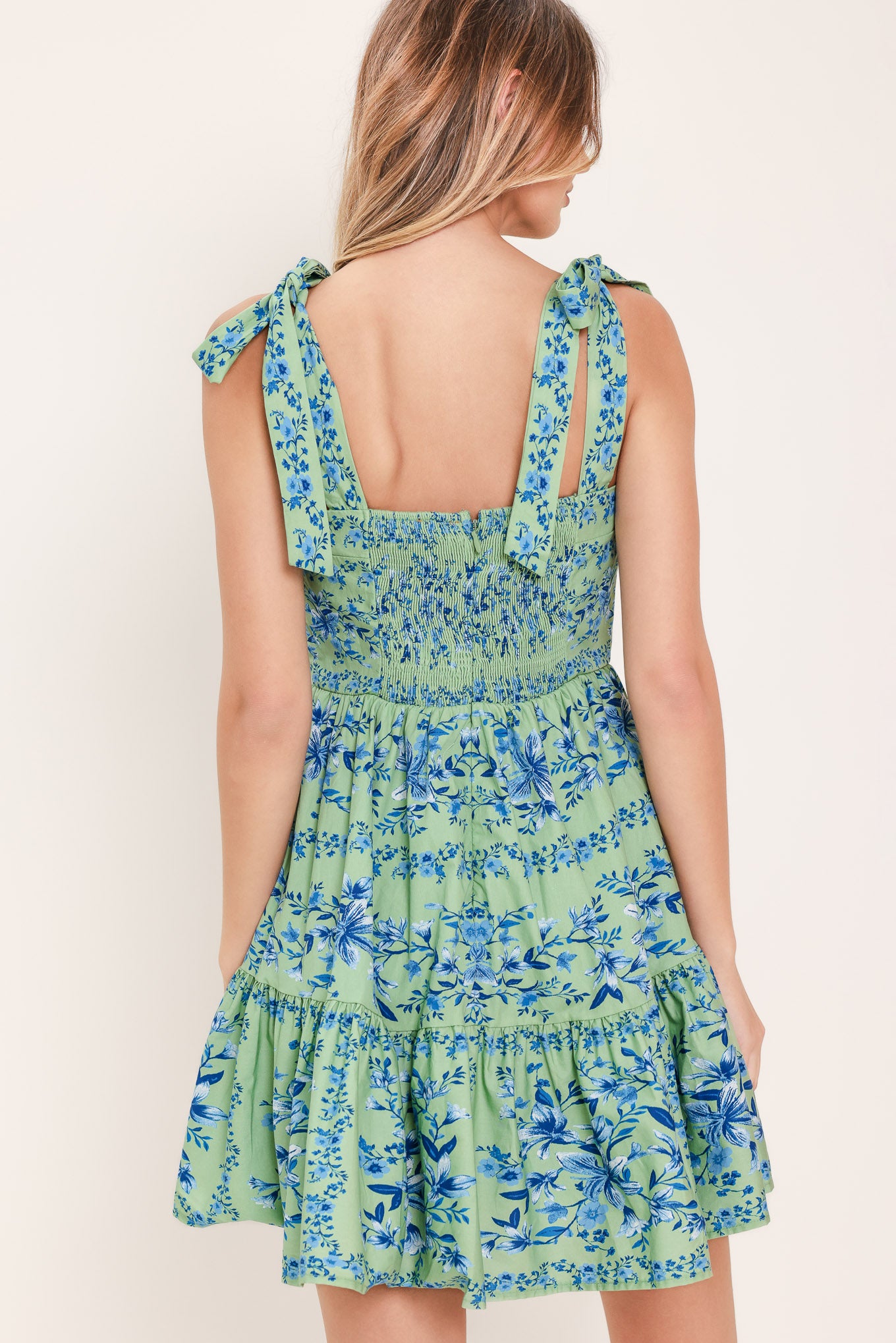 Woman wearing a green and blue floral mini dress featuring straight neckline, shoulder ties, ruffle hem and smocked back bodice with zipper closure | Flying Tomato