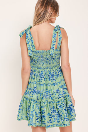 Woman wearing a green and blue floral mini dress featuring straight neckline, shoulder ties, ruffle hem and smocked back bodice with zipper closure | Flying Tomato