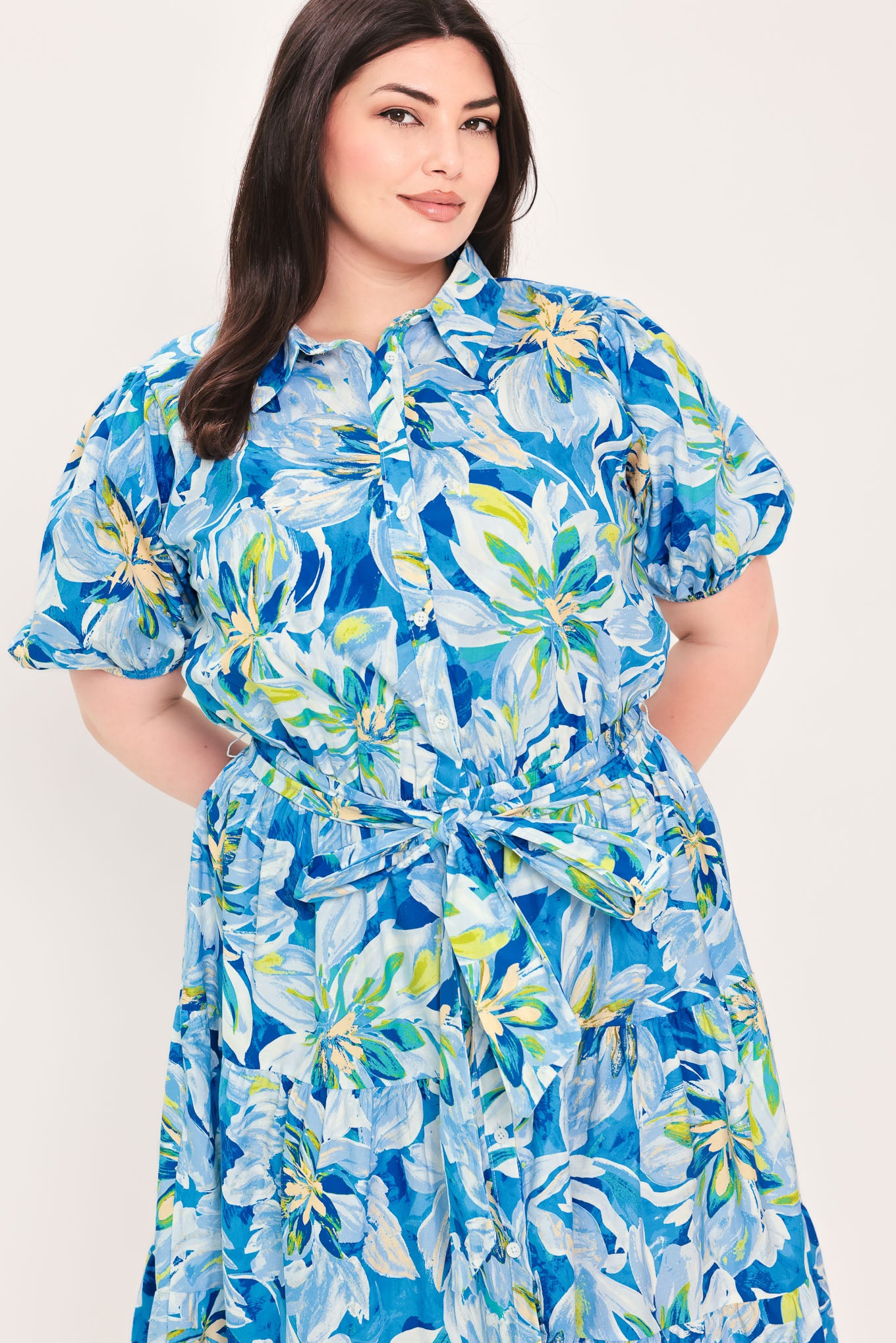 Woman wearing a blue floral midi dress featuring collar, short puff sleeve, button down, self-tie sash, tier skirt and pockets | Flying Tomato