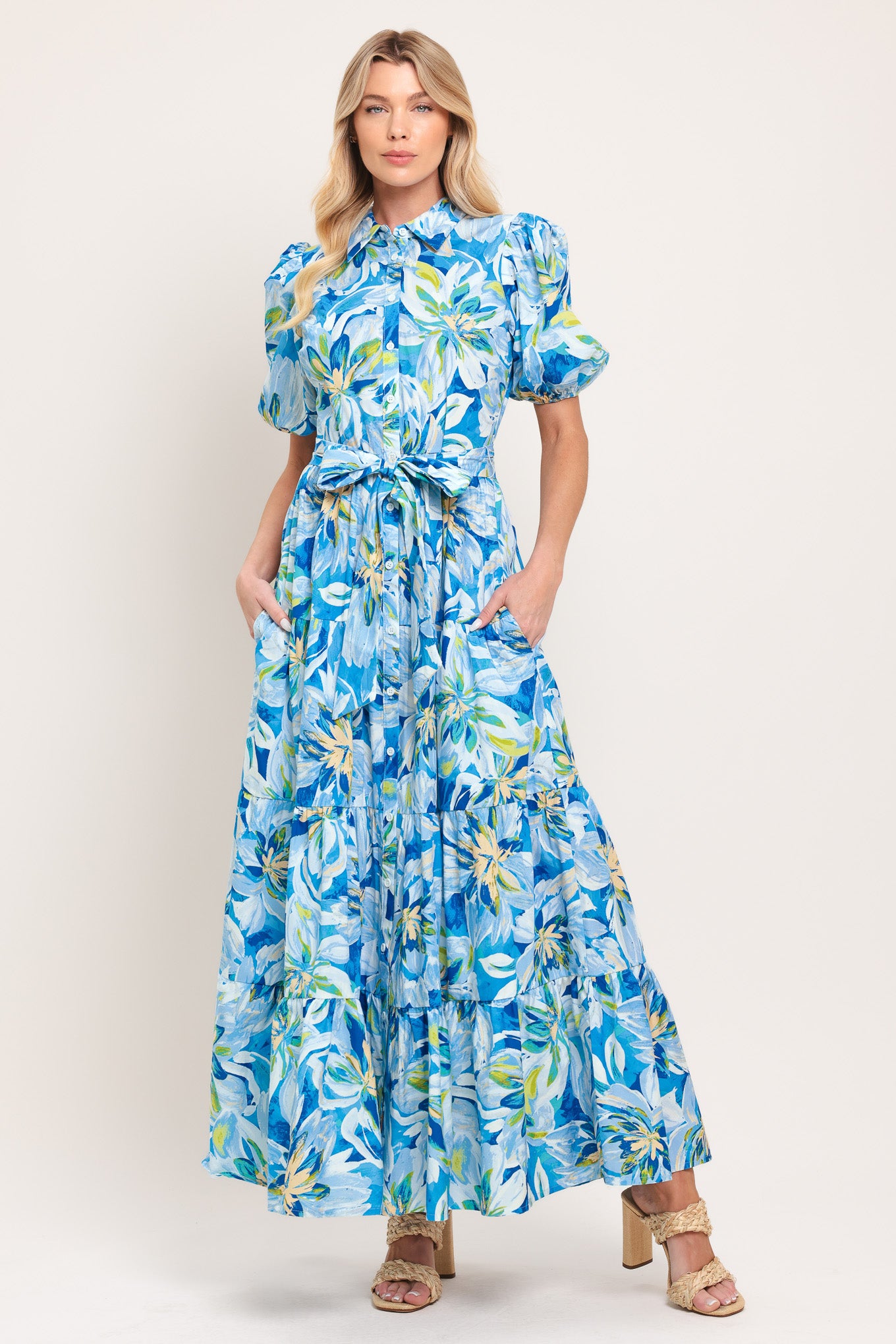 Woman wearing a blue floral midi dress featuring collar, short puff sleeve, button down, self-tie sash, tier skirt and pockets | Flying Tomato
