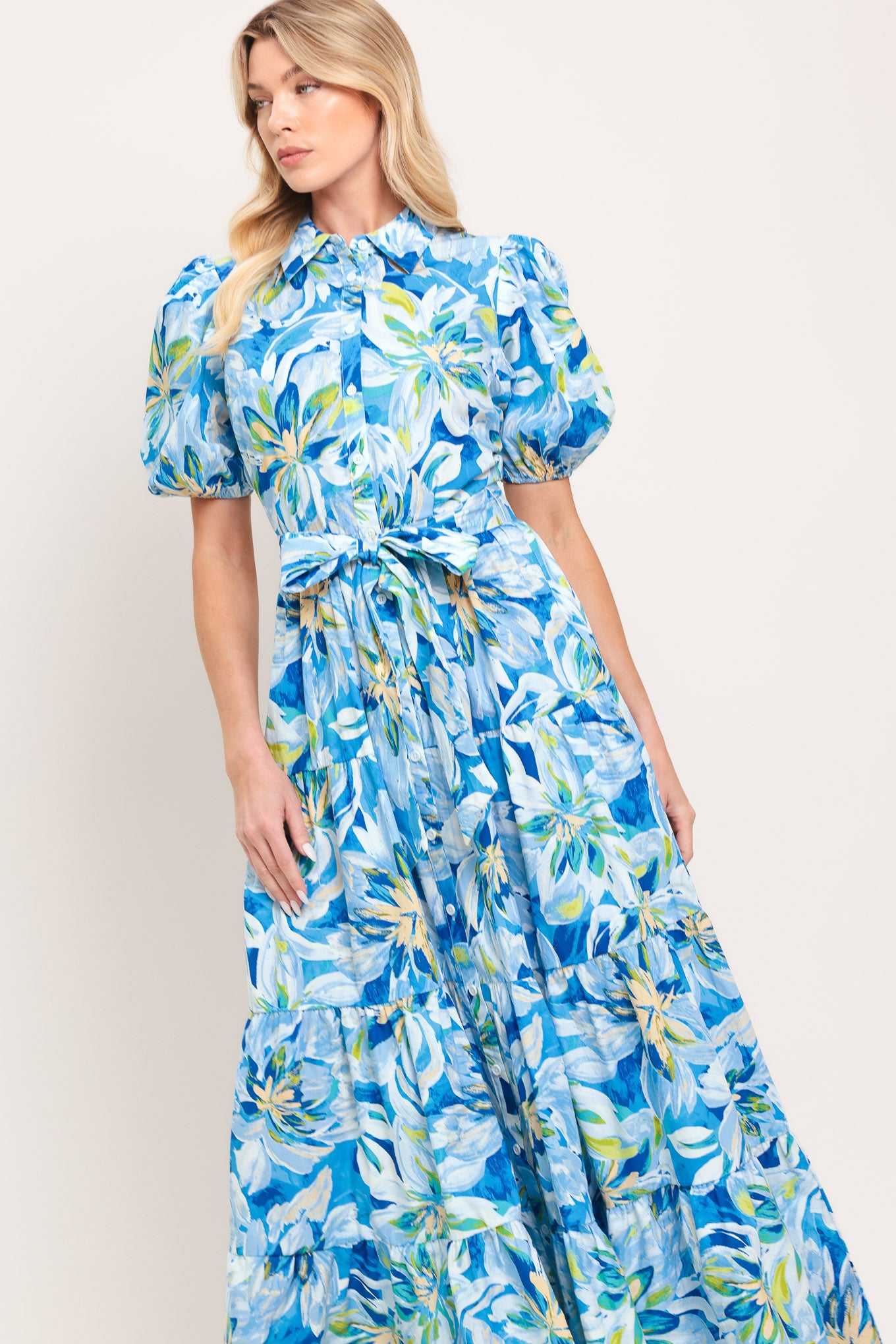 Woman wearing a blue floral midi dress featuring collar, short puff sleeve, button down, self-tie sash, tier skirt and pockets | Flying Tomato