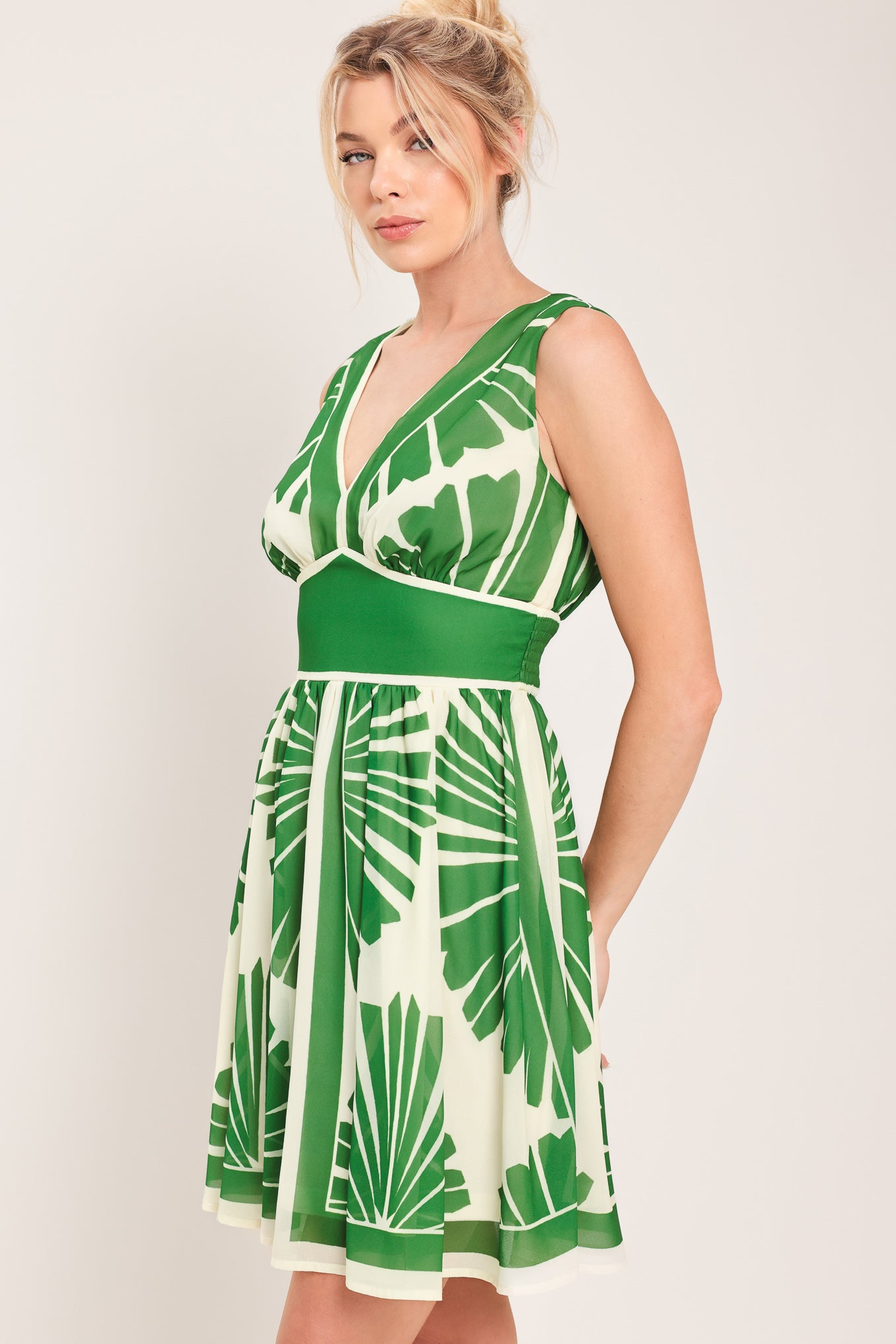 Woman wearing a sleeveless ivory with green leaf pattern mini dress featuring V neckline, full skirt and smocked back waistband | Flying Tomato