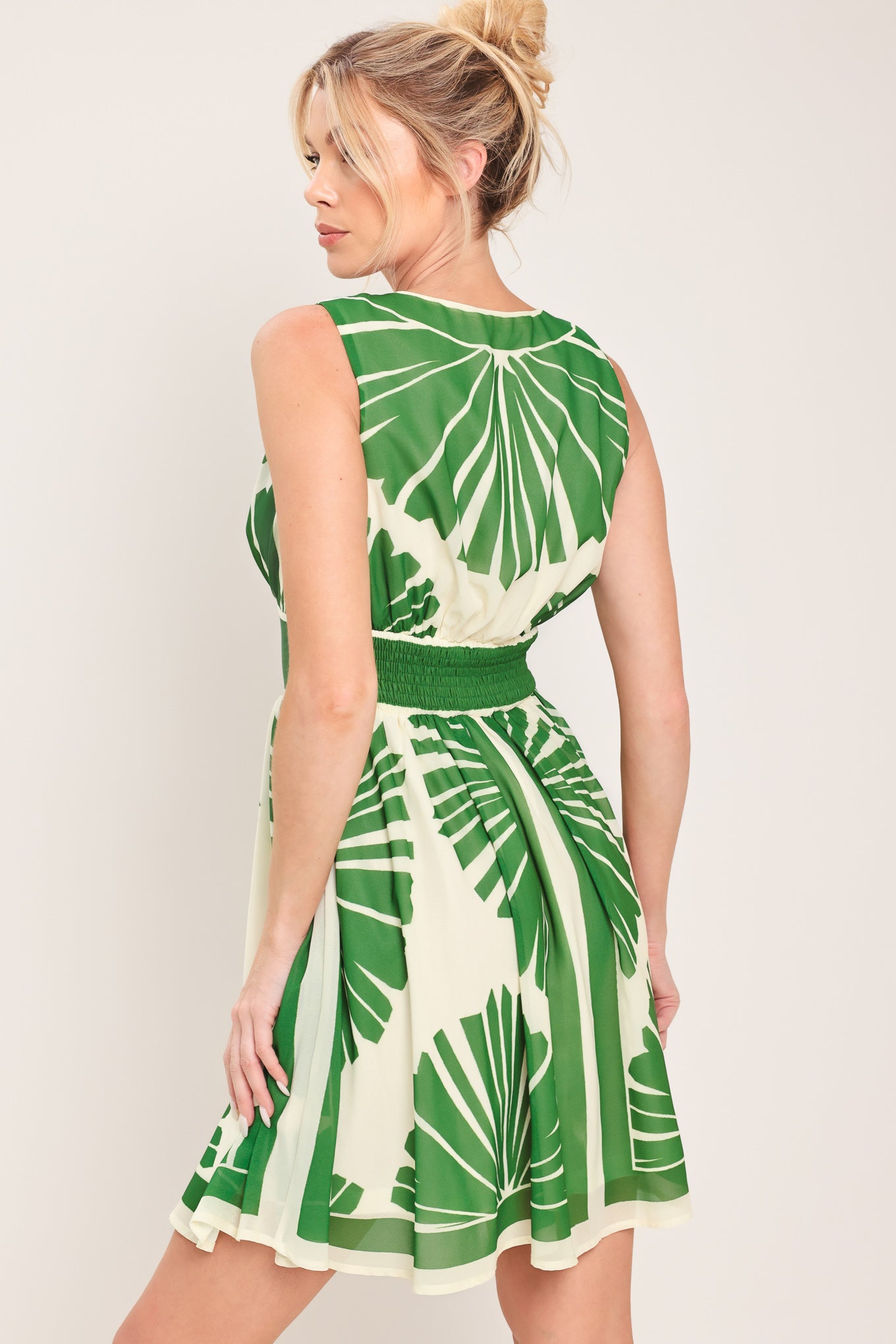 Woman wearing a sleeveless ivory with green leaf pattern mini dress featuring V neckline, full skirt and smocked back waistband | Flying Tomato