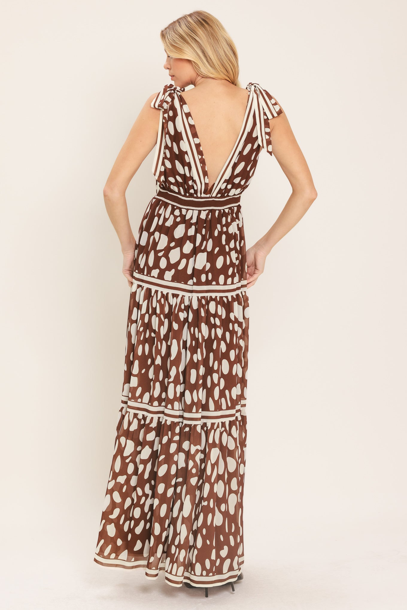 Woman wearing a brown and white spot patterned maxi dress featuring V neckline, shoulder ties, tier skirt and smocked back waistband | Flying Tomato