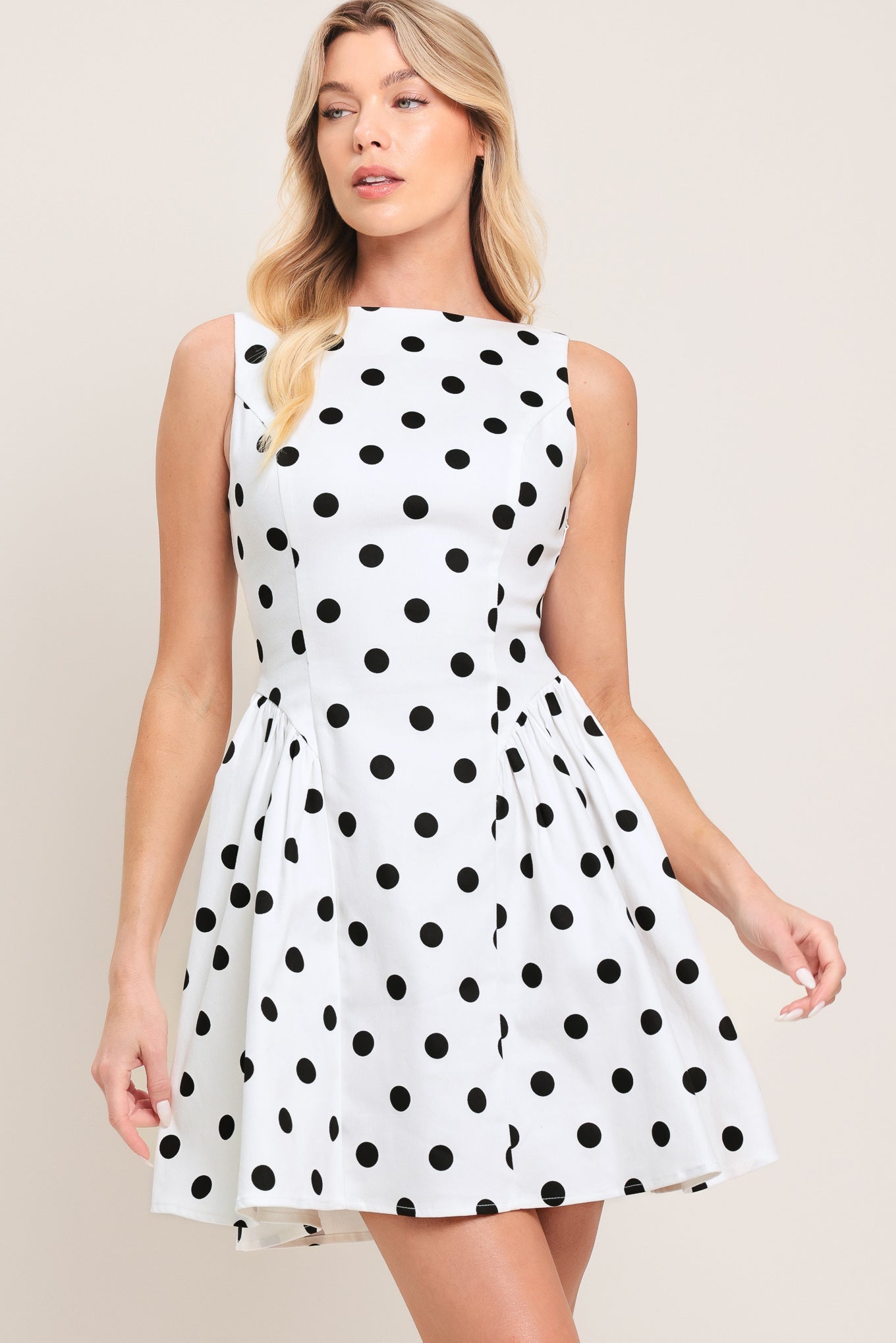 Woman wearing a sleeveless, white mini dress with black polka dots mini dress featuring boat neckline, shirred side panel, and open back with bow detail | Flying Tomato